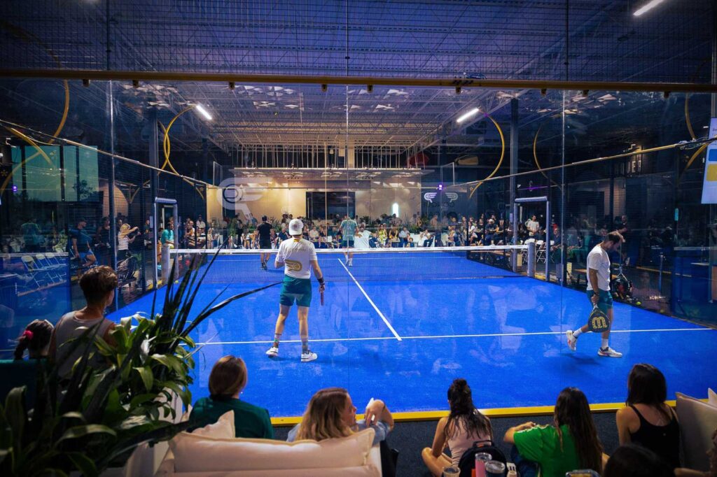 Players competing in an exhibition match at Conquer Padel Tempe while a crowd watches from the lounge area